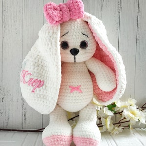 Op de afbeelding: Een witte gehaakte bunny met roze accenten. De bunny heeft een roze strik op zijn hoofd, roze oren en roze voeten. De bunny houdt zijn oren omhoog en heeft een lieve uitdrukking op zijn gezicht. De bunny zit op een wit oppervlak met een witte houten achtergrond.