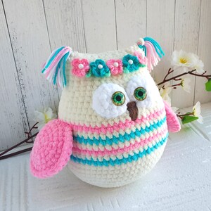 May include: A crocheted owl toy with pink and blue stripes, white fur, green eyes, and a flower crown. The owl has a pink wing and white tassels on its head.