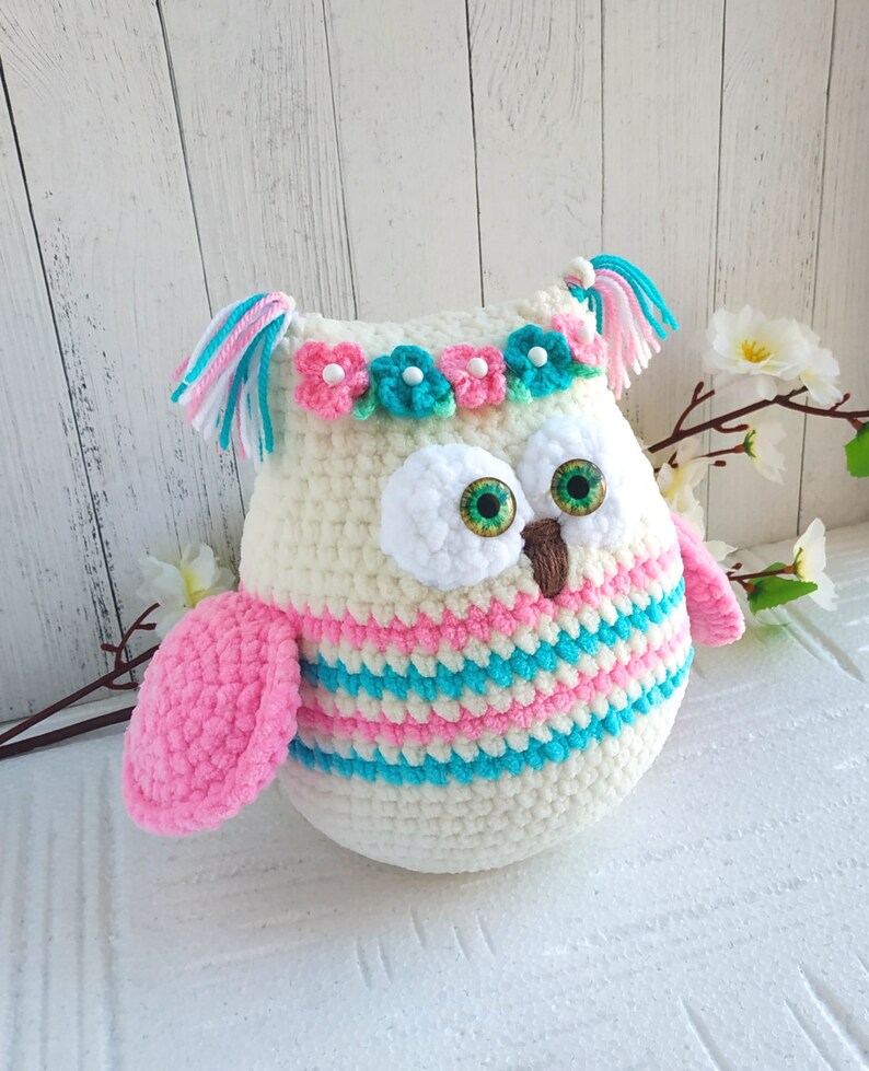 May include: A crocheted owl toy with pink and blue stripes, white fur, green eyes, and a flower crown. The owl has a pink wing and white tassels on its head.