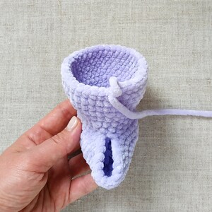 Puede incluir: Una taza de ganchillo lavanda con un agujero en la parte inferior y una larga cola de hilo lavanda unida a la parte superior.