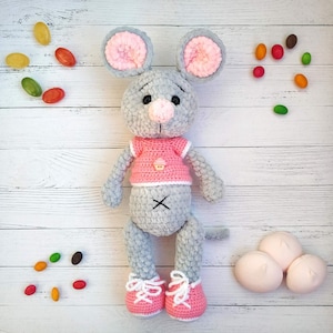 May include: A gray crocheted mouse toy with pink ears, a pink shirt, and white shoes. The mouse is sitting on a white wooden surface with colorful candies scattered around it.