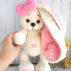 Op de afbeelding: Een witte gehaakte bunny met roze oren en een roze strik. De bunny heeft zwarte ogen en een zwarte neus. De bunny staat op een wit oppervlak.