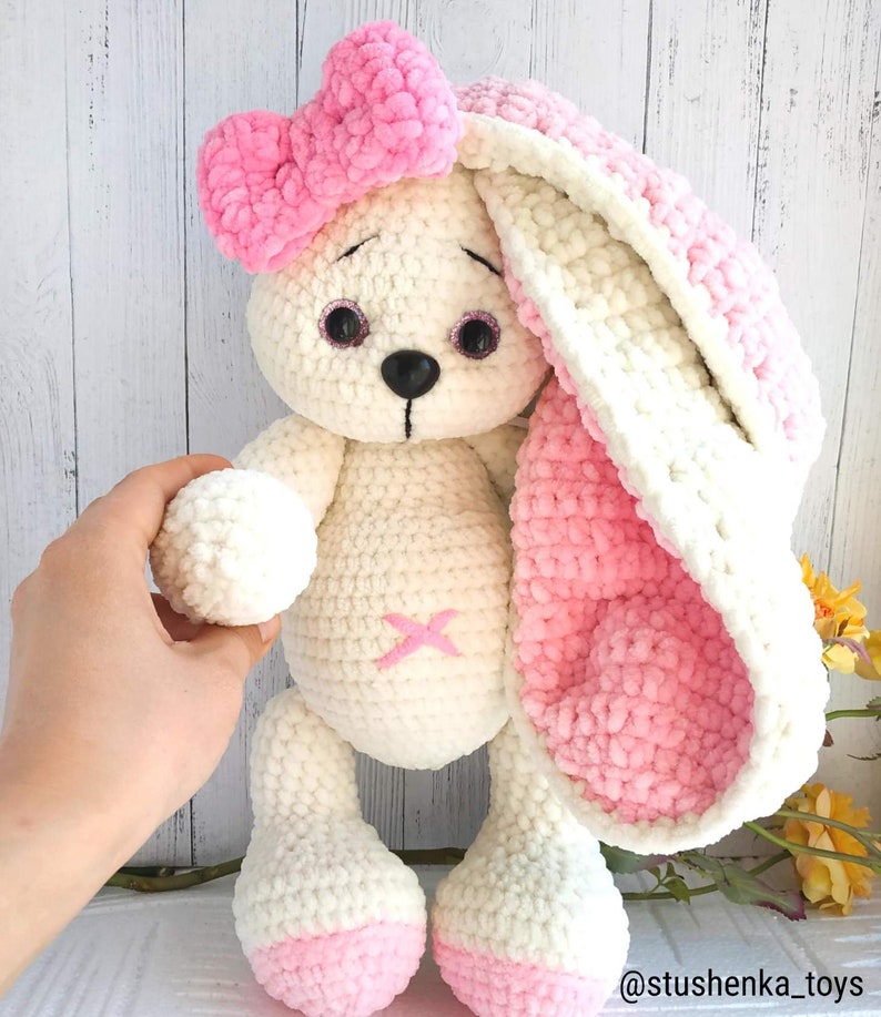 Op de afbeelding: Een witte gehaakte bunny met roze oren en een roze strik. De bunny heeft zwarte ogen en een zwarte neus. De bunny staat op een wit oppervlak.