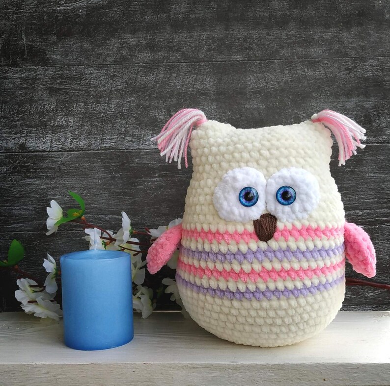 May include: A crocheted owl plush toy with pink and purple stripes, white fur, and blue eyes. The owl is sitting on a white surface.