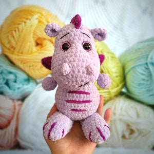 Puede incluir: Un juguete de peluche de drag&oacute;n morado tejido a crochet con rayas rosas y grandes ojos negros. El juguete est&aacute; siendo sostenido por una mano.