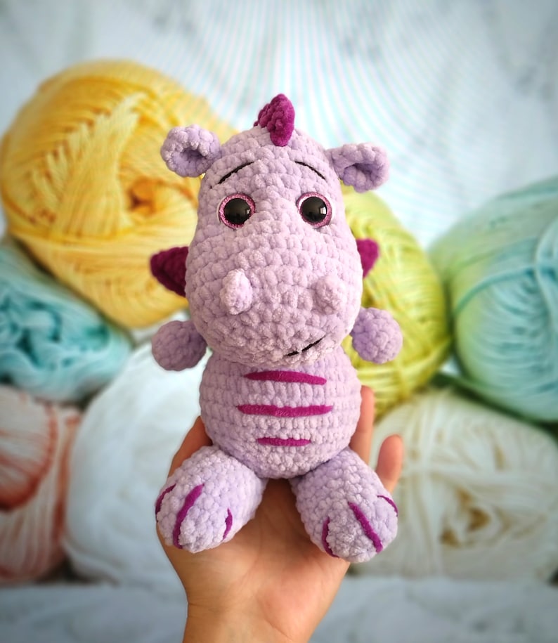 Puede incluir: Un juguete de peluche de drag&oacute;n morado tejido a crochet con rayas rosas y grandes ojos negros. El juguete est&aacute; siendo sostenido por una mano.