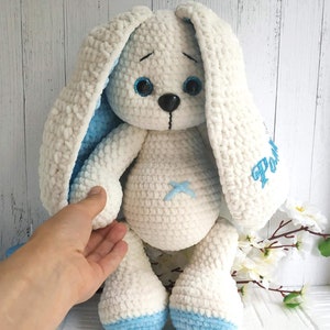 Op de afbeelding: Een witte gehaakte bunny met blauwe oren en voeten. De bunny heeft geborduurde blauwe ogen en een zwarte neus. De bunny zit op een wit houten oppervlak. De bunny heeft de naam "Pom" geborduurd op zijn zijkant.