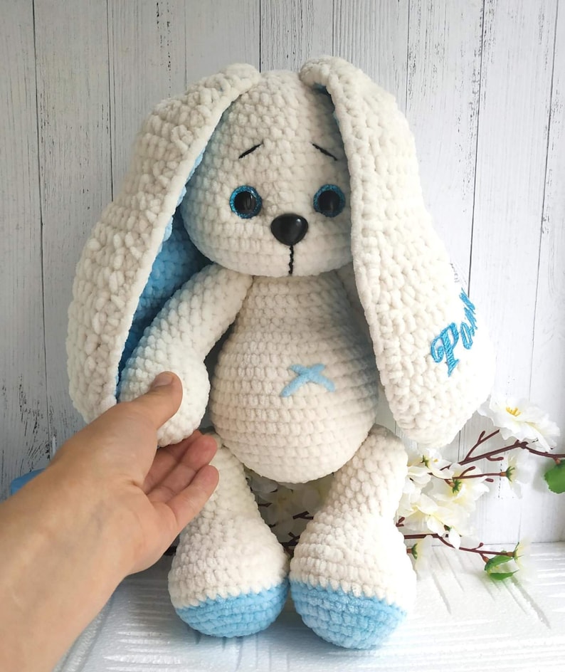 Op de afbeelding: Een witte gehaakte bunny met blauwe oren en voeten. De bunny heeft geborduurde blauwe ogen en een zwarte neus. De bunny zit op een wit houten oppervlak. De bunny heeft de naam "Pom" geborduurd op zijn zijkant.