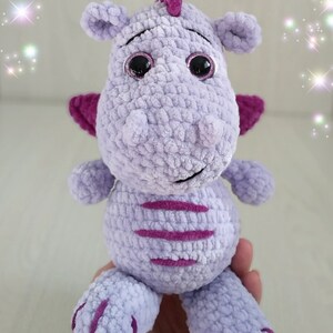 Puede incluir: Un juguete de peluche de drag&oacute;n de crochet morado con detalles en rosa y grandes ojos negros. El juguete est&aacute; sentado en una mano.