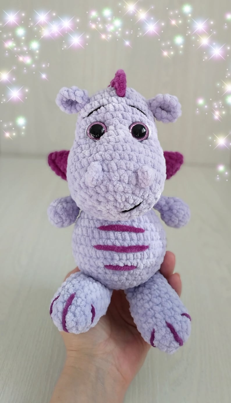 Puede incluir: Un juguete de peluche de drag&oacute;n de crochet morado con detalles en rosa y grandes ojos negros. El juguete est&aacute; sentado en una mano.