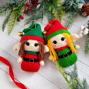 Puede incluir: Dos adornos de elfo de crochet con sombreros rojos y verdes, caras blancas y cabello amarillo. El elfo de la izquierda lleva un atuendo rojo y verde, mientras que el elfo de la derecha lleva un atuendo verde y blanco.