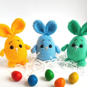 May include: Three crocheted Easter bunny toys, one yellow, one blue, and one green, with black eyes and white noses. The bunnies are sitting on a white surface with colorful Easter eggs.