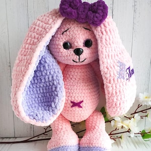 Op de afbeelding: Een roze gehaakte konijn met paarse oren en een paarse strik. Het konijn heeft zwarte ogen en een zwarte neus. Het konijn zit op een wit oppervlak met een tak met witte bloemen erachter.