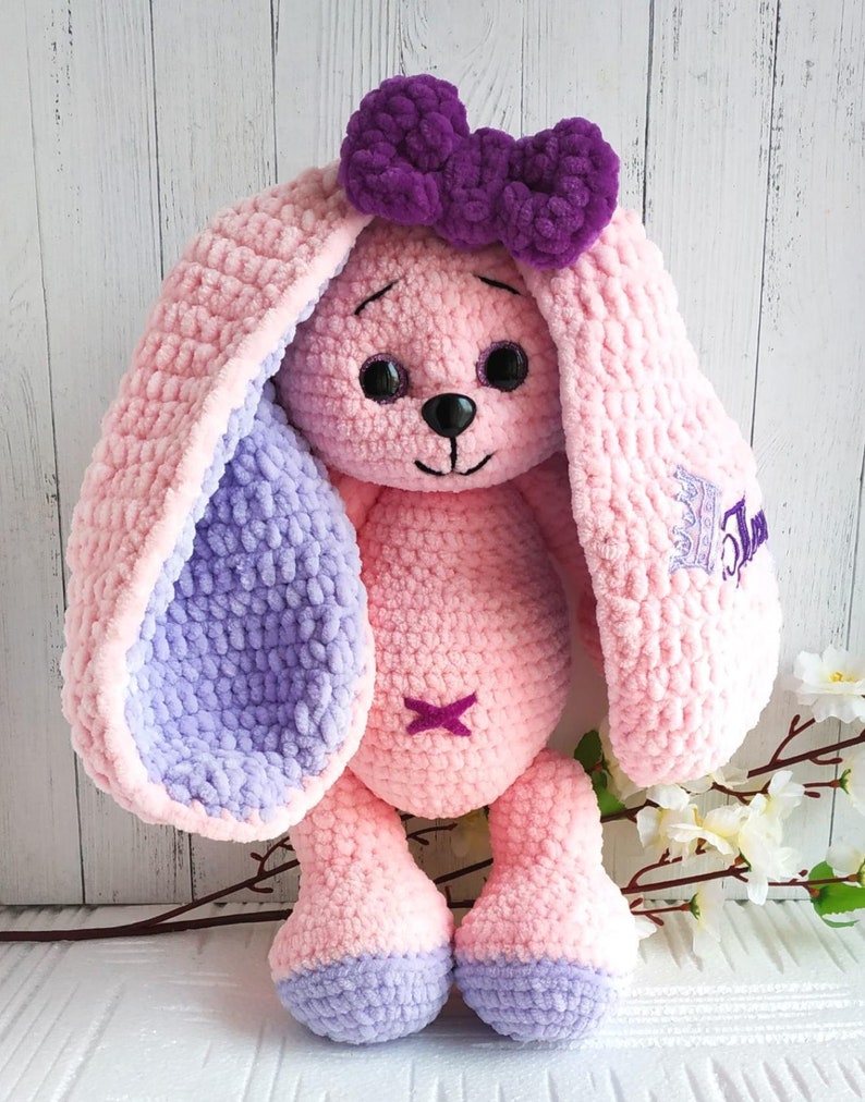 Op de afbeelding: Een roze gehaakte konijn met paarse oren en een paarse strik. Het konijn heeft zwarte ogen en een zwarte neus. Het konijn zit op een wit oppervlak met een tak met witte bloemen erachter.