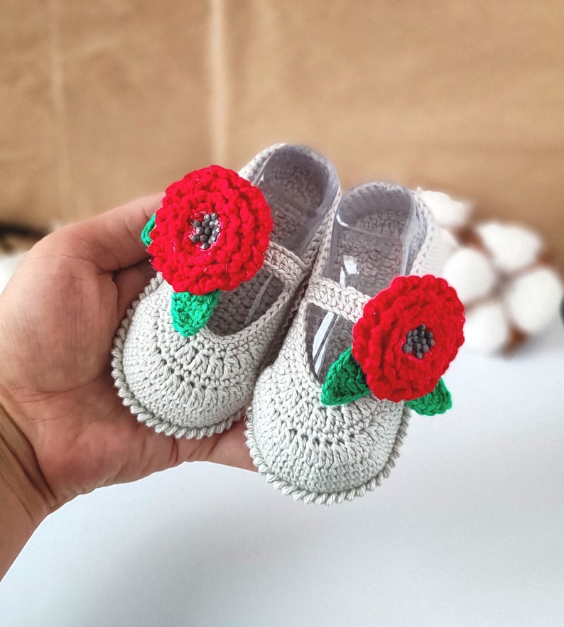 Pu&ograve; includere: Un paio di scarpine per neonati in crochet grigio con decorazioni floreali rosse. Ogni scarpetta ha un fiore rosso con foglie verdi.