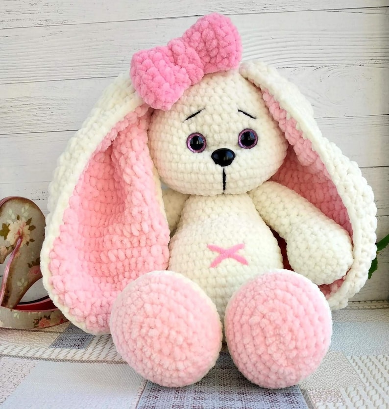 Op de afbeelding: Een witte gehaakte bunny met roze oren en een roze strik. De bunny heeft zwarte ogen en een zwarte neus. De bunny zit op een wit oppervlak.