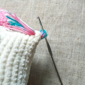 May include: A close-up of a white crocheted cushion with a pink, white and blue yarn tail being sewn on with a crochet hook.