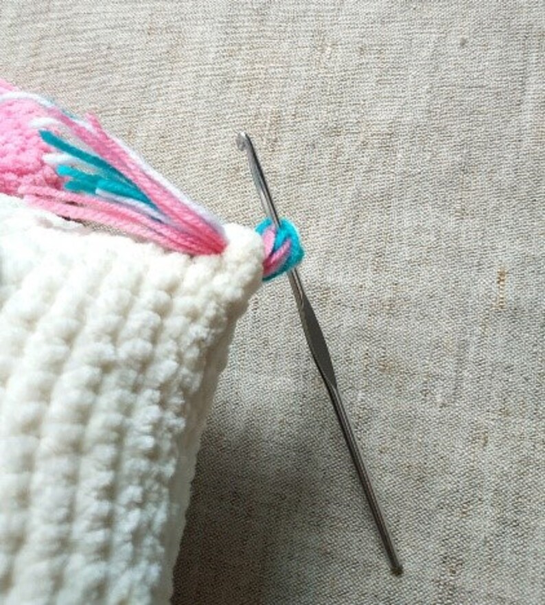 May include: A close-up of a white crocheted cushion with a pink, white and blue yarn tail being sewn on with a crochet hook.