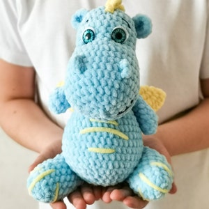 Puede incluir: Un juguete de peluche de drag&oacute;n de color azul claro tejido a crochet con detalles amarillos y ojos verdes. El drag&oacute;n est&aacute; sentado en las manos de una persona.