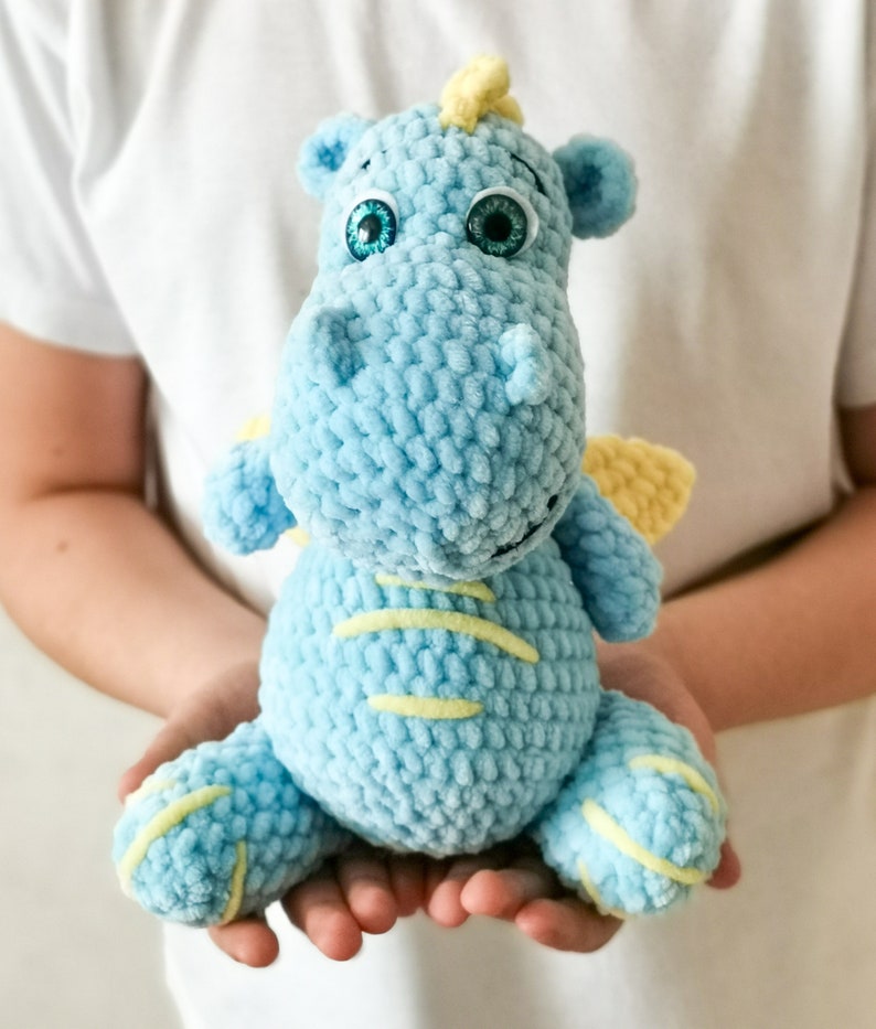 Puede incluir: Un juguete de peluche de drag&oacute;n de color azul claro tejido a crochet con detalles amarillos y ojos verdes. El drag&oacute;n est&aacute; sentado en las manos de una persona.