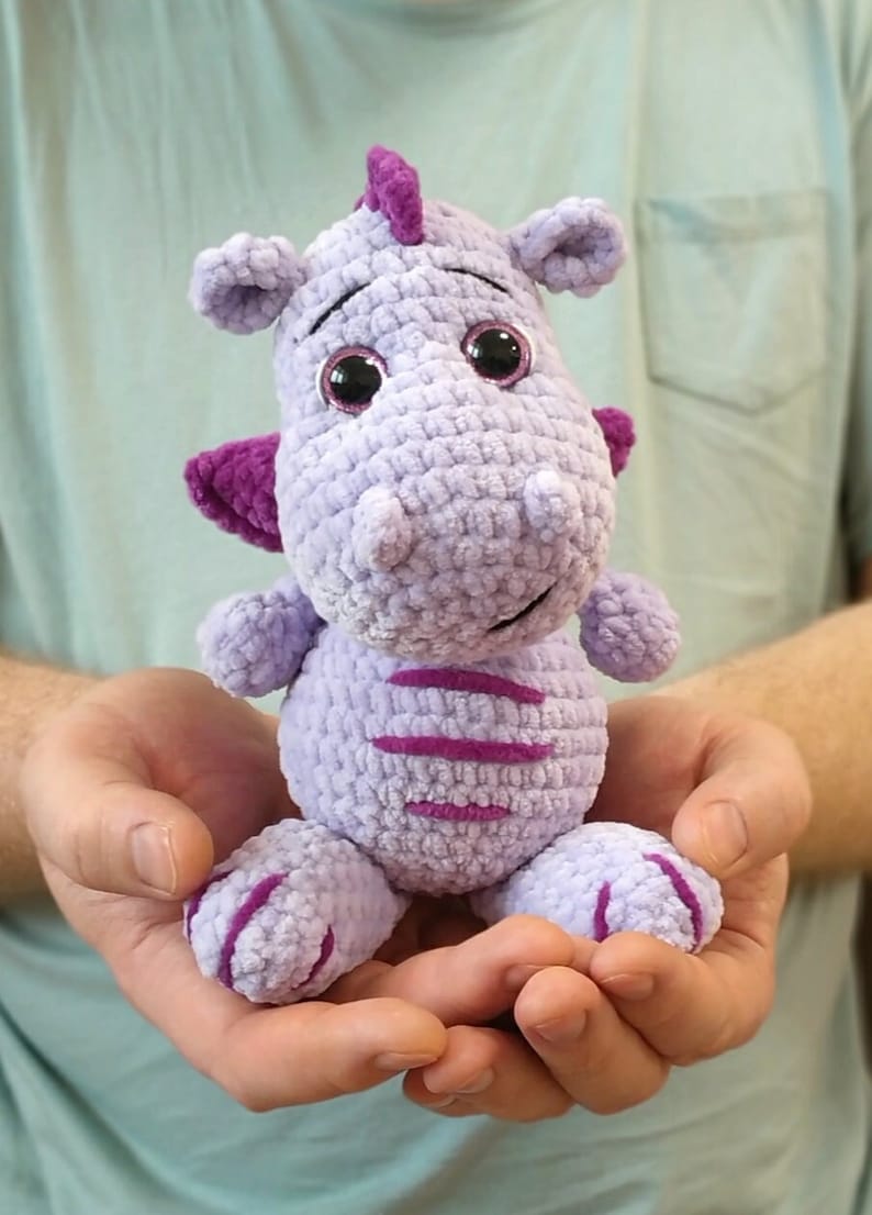 Puede incluir: Un juguete de peluche de drag&oacute;n de color morado tejido a crochet con detalles en rosa y grandes ojos negros. El drag&oacute;n est&aacute; sentado en las palmas de las manos de una persona.