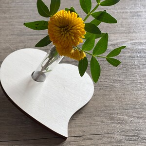 Könnte beinhalten: Eine weiße herzförmige Holzvase mit einem durchsichtigen Glasrohr für Blumen. Ein einziger Stiel gelber Chrysanthemen steht in der Vase.