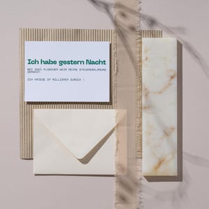Può includere: Una composizione di natura morta con una carta bianca con testo verde, una busta color crema, un blocco di marmo rettangolare e cartone ondulato. Il testo tedesco sulla carta recita: "Ich habe gestern Nacht..." su sfondo rosa chiaro.