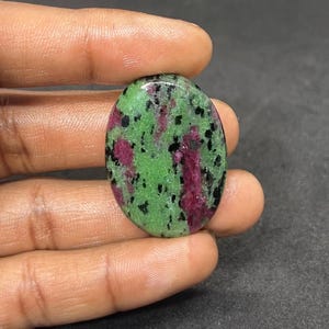 May include: An oval gemstone with a striking colour palette. The stone displays a base of vivid green, interspersed with black specks and areas of rich red. The polished surface catches the light, accentuating the natural patterns.