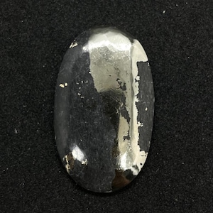 Pode incluir: Objeto metálico oval com uma superfície escura e reflexiva. A superfície tem áreas onde o revestimento está lascado, revelando uma cor mais clara por baixo. O objeto está sobre um fundo escuro e texturizado.