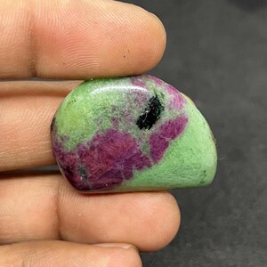 Pode incluir: Uma pedra preciosa polida com formato arredondado. A pedra apresenta uma mistura de cores, incluindo verde, magenta escuro e preto. O verde é a cor dominante, com manchas magenta e pretas. A pedra é segurada entre dois dedos.