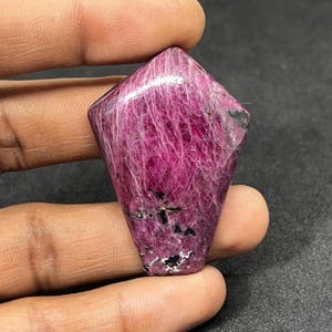 May include: A faceted, deep magenta gemstone with a polished surface. The stone has a unique shape, with a flat top and a tapered bottom. Veins of lighter pink and black inclusions are visible throughout the stone.