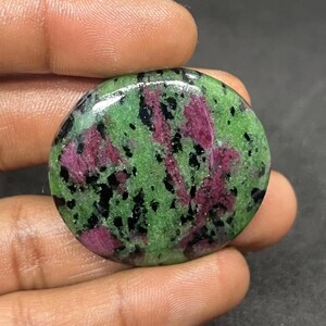 May include: A polished, round gemstone featuring a vibrant mix of colors. The stone displays a speckled pattern of deep green, interspersed with streaks of magenta and black. The gemstone is held in a hand.