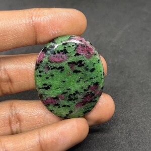 May include: Oval-shaped zoisite gemstone with a vibrant green base, speckled with black and patches of deep red. The polished stone is held between fingers, showcasing its natural colors and textures.