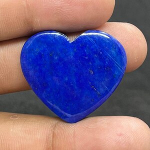Puede incluir: Una gema de lapislázuli pulida en forma de corazón. La piedra azul oscuro tiene vetas sutiles y una superficie lisa y reflectante. La gema se sostiene entre dos dedos, mostrando su forma y color.