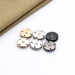 May include: Six metal snap fasteners in gold, silver, and black. Each fastener is round with a central hole and four small rectangular cutouts. A tan fabric is in the background.