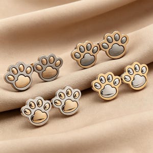 Könnte beinhalten: Sechs Manschettenknöpfe mit Pfotenabdruck in Gold- und Silbertönen. Die Manschettenknöpfe sind paarweise angeordnet, jedes mit einem detaillierten Pfotenabdruck-Design. Der Hintergrund ist ein neutraler beigefarbener Stoff.
