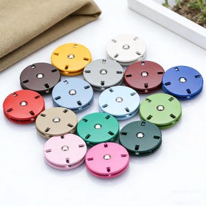 Puede incluir: Una colección de coloridos cierres de botón redondos en varios tonos, incluyendo rojo, azul, verde y rosa. Cada cierre tiene un centro plateado y un diseño circular con ranuras. Los cierres están dispuestos en un grupo, mostrando su diversa paleta de colores.