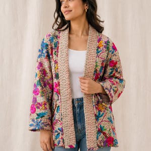 Op de afbeelding: Beige jas met bloemenprint en een gewatteerd ontwerp. De jas heeft een levendig patroon van roze, blauwe en gele bloemen. De jas is open en onthult een witte top en een blauwe jeans.