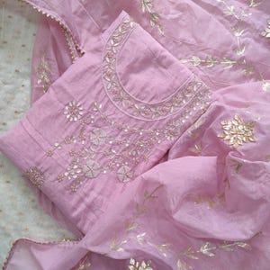 Op de afbeelding: Een lichtroze geborduurde salwar kameez set. De set bevat een tuniek en een bijpassende sjaal, beide versierd met ingewikkelde goudkleurige bloemen- en bladborduurwerken. De tuniek heeft gedetailleerde borduursels rond de halslijn en aan de voorkant.