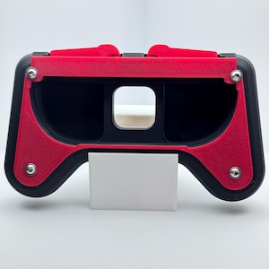 May include: A black and red VR headset with a rectangular opening for the phone. The headset has a red top and sides, with silver screws. A white rectangular piece is at the bottom.