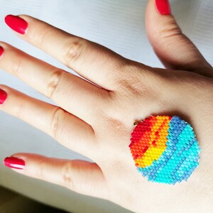 Peut inclure: Un motif de soleil arc-en-ciel color&eacute; en perles sur la main d'une personne. Le soleil est fait de petites perles color&eacute;es dans des tons de rouge, orange, jaune, vert, bleu et violet. Les perles sont dispos&eacute;es en cercle, cr&eacute;ant un effet de soleil.