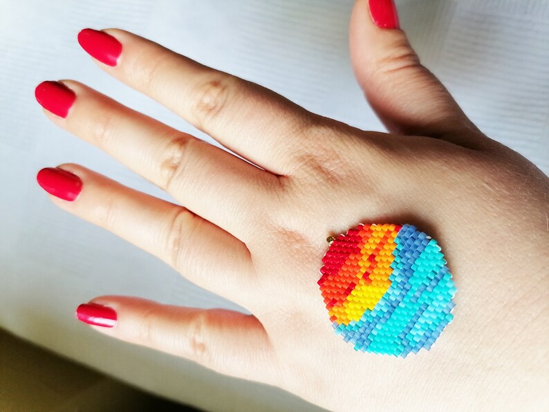 Peut inclure: Un motif de soleil arc-en-ciel color&eacute; en perles sur la main d'une personne. Le soleil est fait de petites perles color&eacute;es dans des tons de rouge, orange, jaune, vert, bleu et violet. Les perles sont dispos&eacute;es en cercle, cr&eacute;ant un effet de soleil.