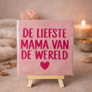 May include: A pink square card with the text "DE LIEFSTE MAMA VAN DE WERELD" and a heart symbol, displayed on a small wooden easel. The card is in a clear plastic sleeve. A lit candle and other objects are in the background.