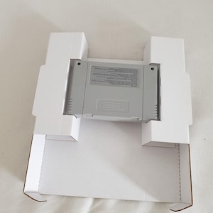 Könnte beinhalten: Eine graue, rechteckige Videospielkassette mit einem Etikett, das das Nintendo-Logo und Text zeigt, sicher platziert in einem weißen Kartoneinsatz. Die Kassette hat eine gerippte Rückseite und wird von weißen, gefalteten Laschen gehalten.