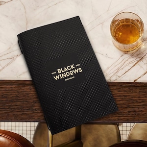 以下が含まれることがあります： 「BLACK WINDOWS RESTAURANT」と金色の文字が入った黒いメニューが、琥珀色の液体が入ったグラスの隣のマーブルテーブルに置かれています。テーブルは濃い木材のトリムで、茶色の革張りのバースツールの近くにあります。