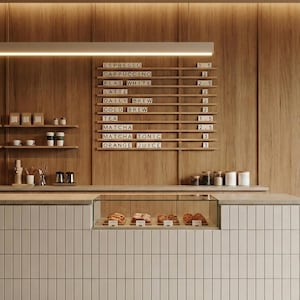 Puede incluir: Interior de una cafetería con un fondo de madera y una pizarra de menú que enumera opciones de café y té. Una vitrina muestra pasteles. El mostrador está alicatado en blanco y el suelo tiene un patrón moteado.