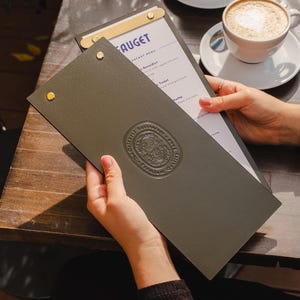 May include: A person holding an open menu at a cafe. The menu is dark green with gold accents and a debossed emblem. A cup of coffee with latte art sits on a white saucer nearby. The cafe table is made of wood.