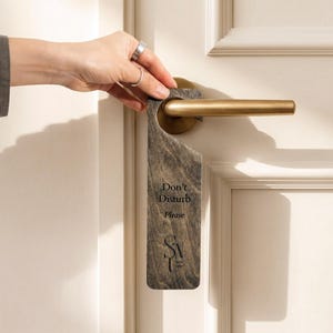 Pode incluir: Um pendurador de porta de madeira cinzento-escuro com uma textura de grão de madeira visível. O texto "Don't Disturb Please" está impresso a preto, seguido de um logótipo estilizado na parte inferior. O pendurador está suspenso numa maçaneta.