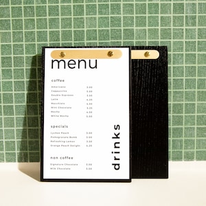 Puede incluir: Un menú negro con interior blanco, con una lista de cafés y bebidas especiales. El menú se sujeta con cierres dorados. La palabra "menú" está en la parte superior, y "drinks" está escrito verticalmente a la derecha.