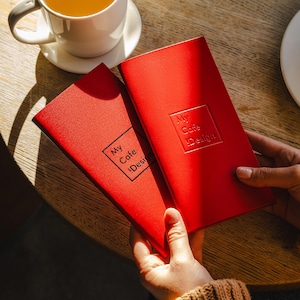 May include: Two red notebooks with the text "My Cafe Design" are held in front of a wooden table. A white teacup and saucer with tea and a chocolate muffin are also on the table. The notebooks have a square logo.